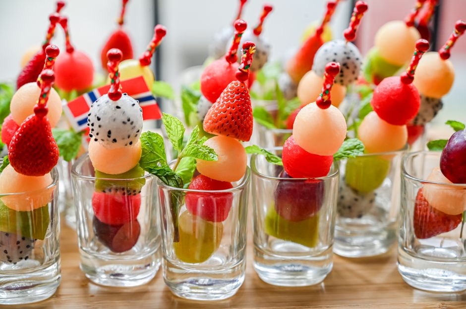 Brochettes colorées de fruits variés dans des verres, décorées de feuilles de menthe et de piques amusants.