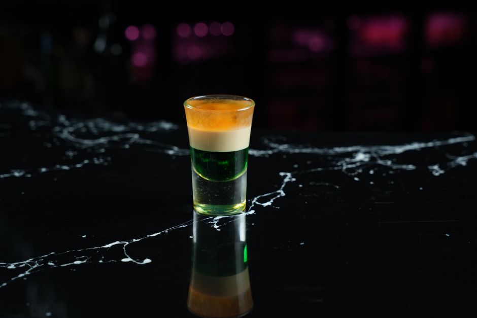 Verre de shot avec trois couches de couleurs : vert, jaune et orange, sur un fond noir marbré.