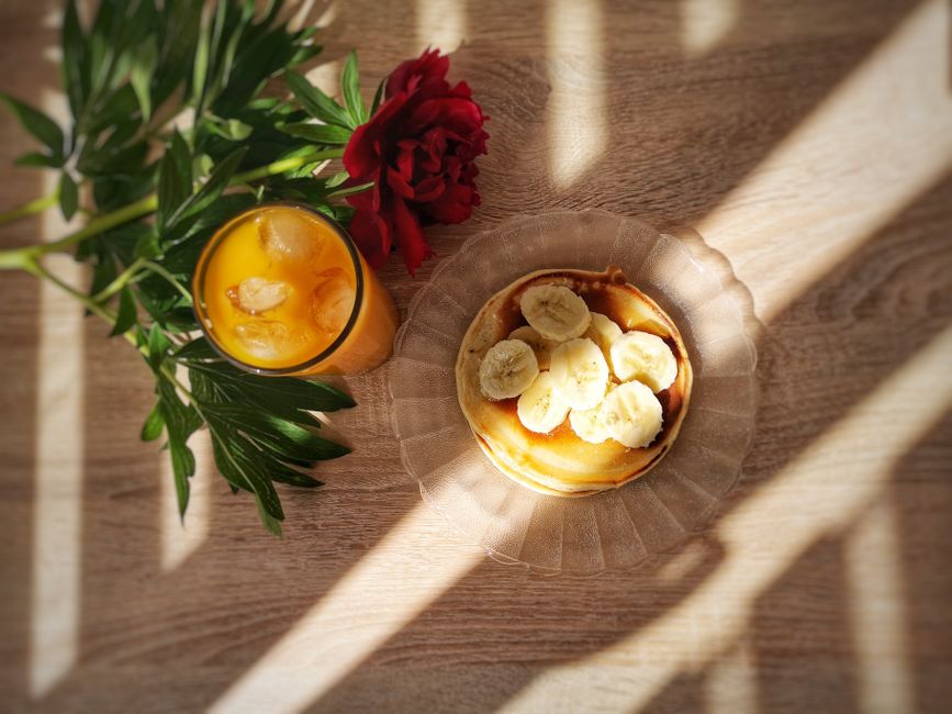 Pancake épais garni de bananes, accompagné d'un verre de jus d'orange et d'une fleur sur une table en bois.