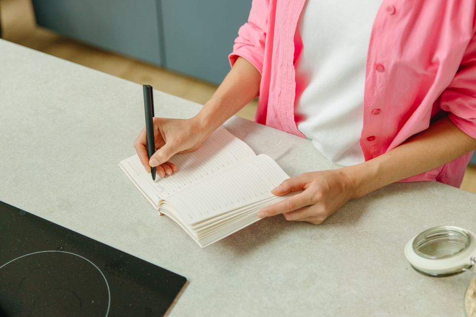 Femme en chemise rose écrivant une liste de courses dans un carnet sur un plan de travail.