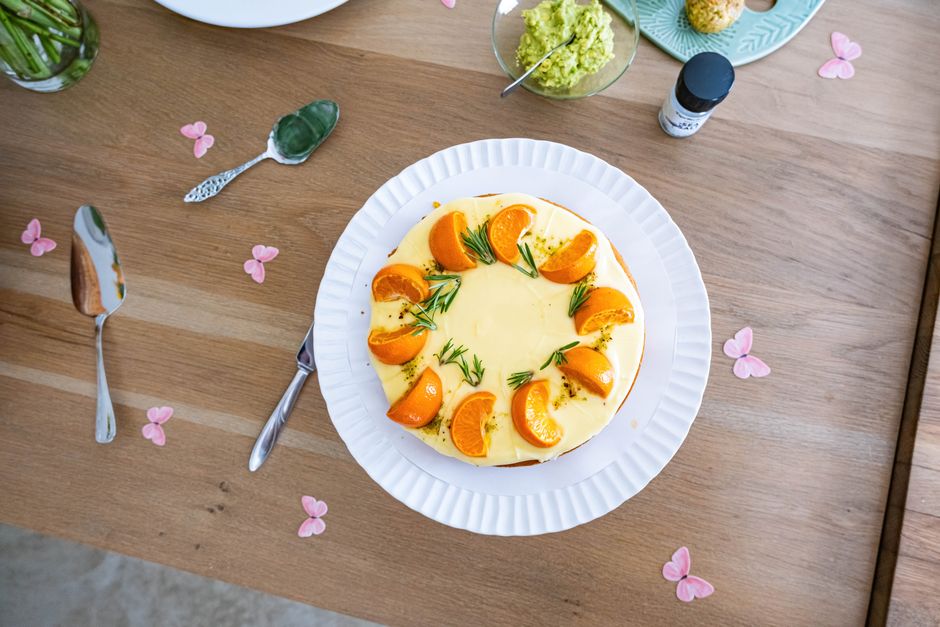 Gâteau salé décoré d'orange et de romarin, sur une assiette blanche, entouré de fleurs en papier sur une table en bois.