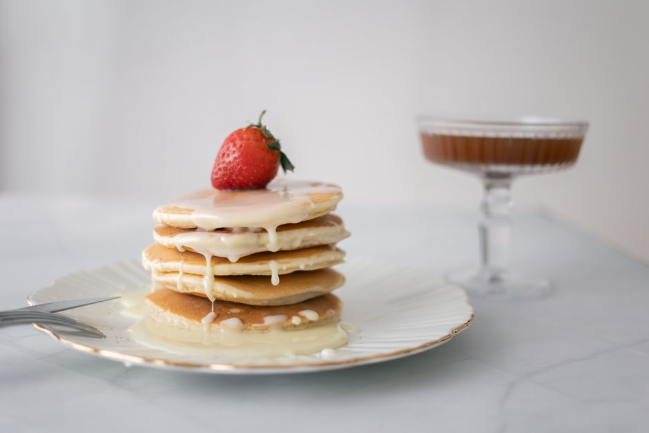 Pancakes épais empilés sur une assiette avec un coulis et une fraise sur le dessus, accompagnés d'un verre de sirop.