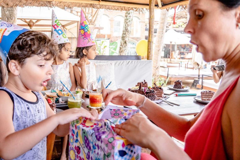 Enfant ouvrant un cadeau d'anniversaire, entouré de camarades portant des chapeaux de fête, avec des gâteaux en arrière-plan.