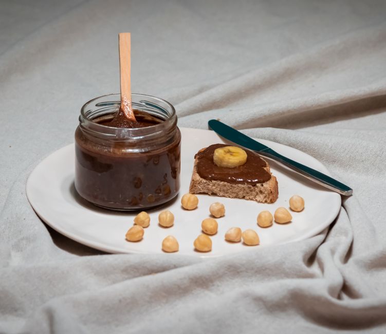 Pâte à tartiner noisette maison dans un pot, tartinée sur une tranche de pain avec des noisettes et une banane.