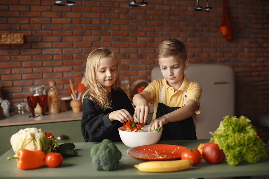 Deux enfants préparent une salade dans une cuisine, entourés de légumes frais sur la table.