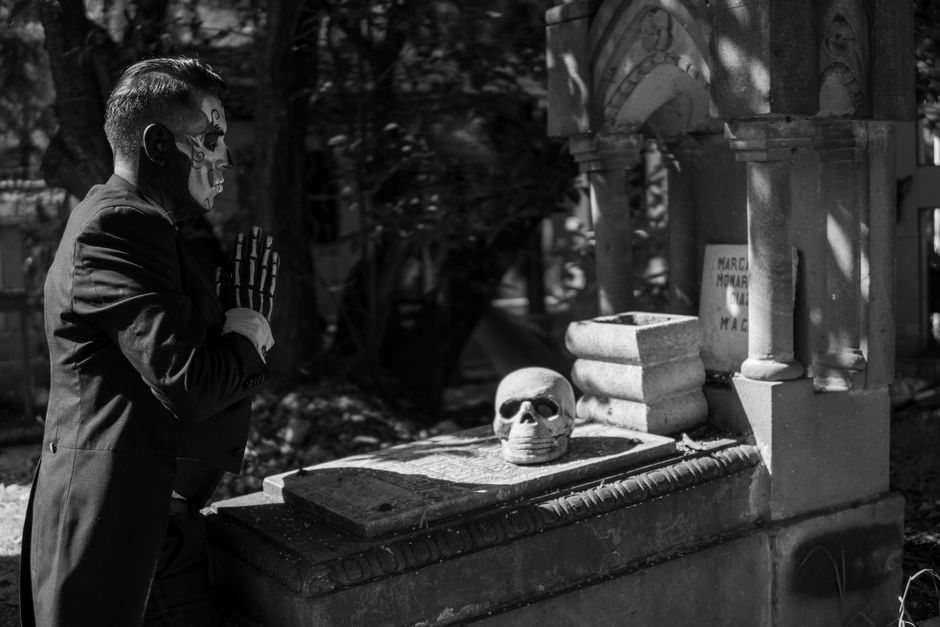 Homme en costume noir avec maquillage de squelette devant une tombe et un crâne en pierre.