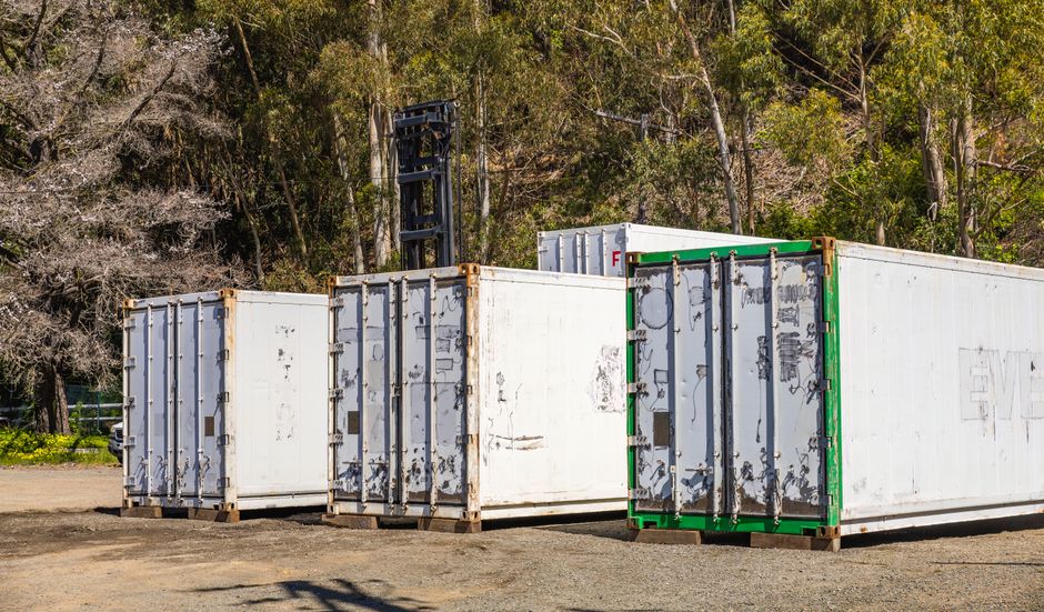 Trois conteneurs de stockage blancs et verts alignés dans un espace extérieur, entourés d'arbres.