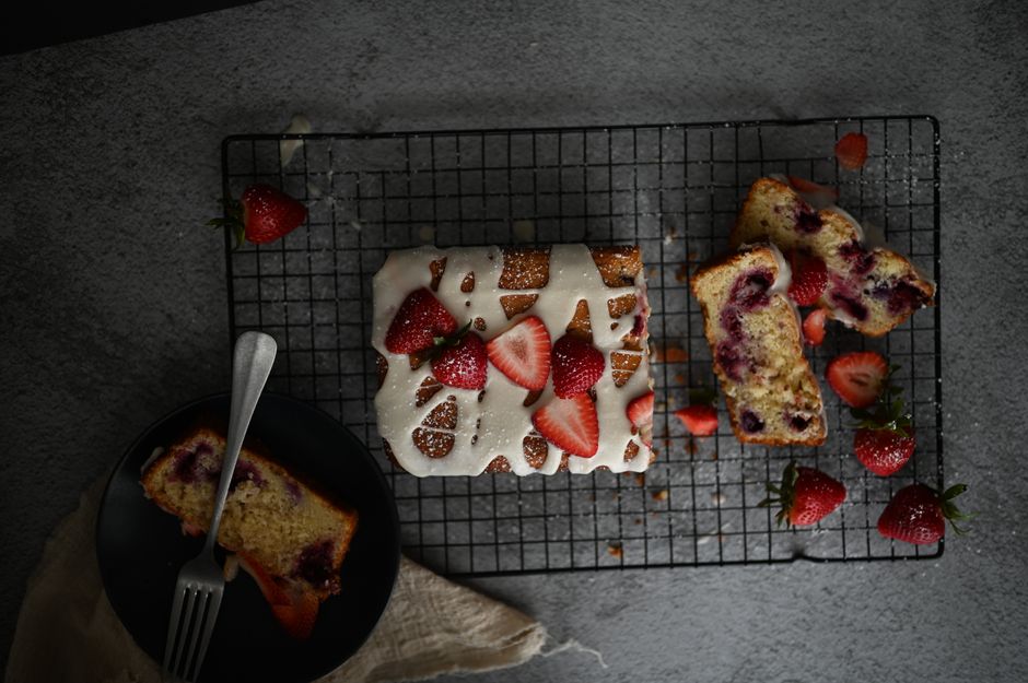 Gâteau au yaourt sur une grille, garni de fraises et de glaçage, avec une part sur une assiette noire.