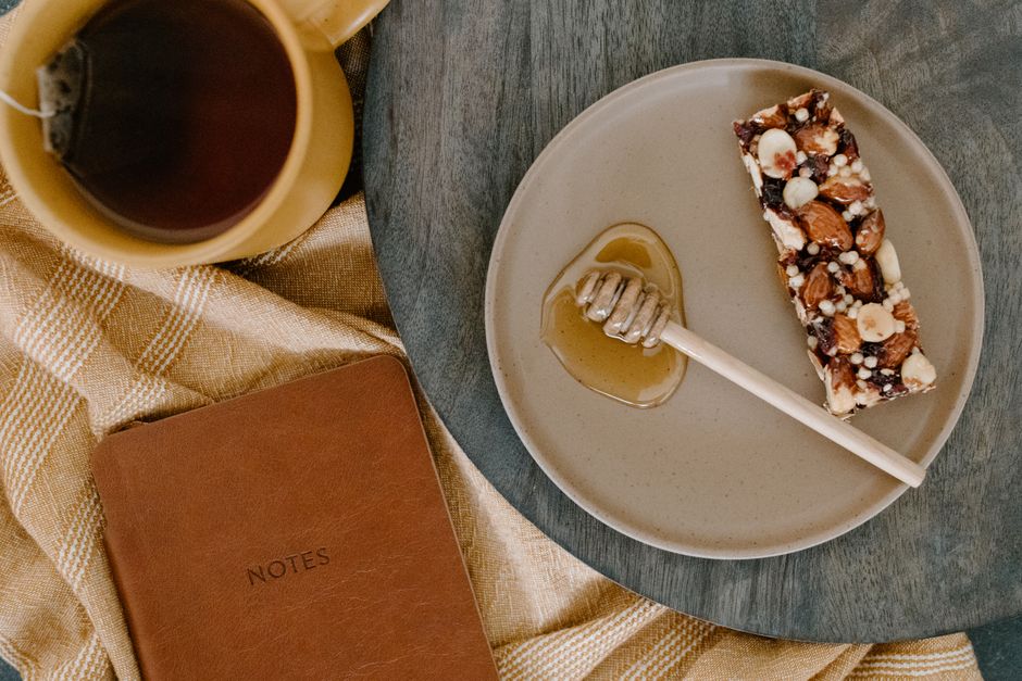 Barre de céréales maison sur une assiette avec du miel, une tasse de thé et un carnet en cuir sur une nappe.