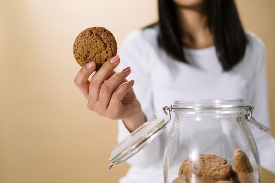 Femme tenant un biscuit près d'un bocal en verre rempli de biscuits sur un fond neutre.